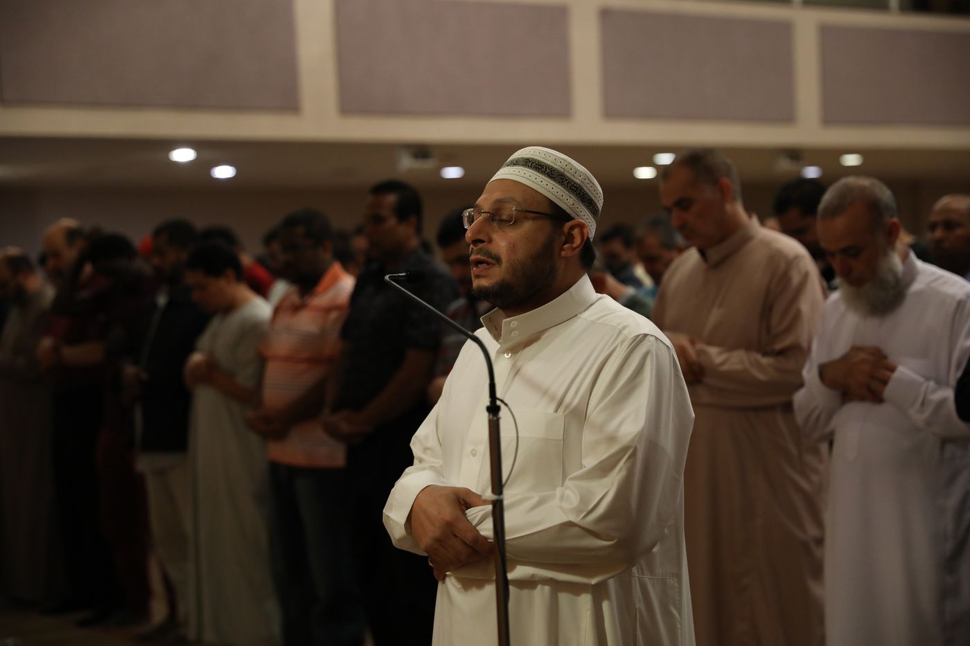 An imam in a white thobe and kufi leading the congregation in prayer during Ramadan